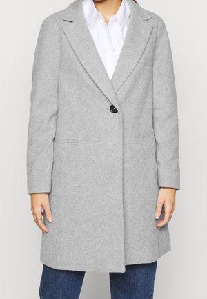Persona che indossa un cappotto di lana grigio chiaro con un solo bottone nero sopra una camicia bianca e jeans blu, mani rilassate ai lati.