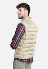 Chaleco acolchado beige con secciones cosidas verticalmente, combinado con una camisa de cuadro en azul marino y rojo. El chaleco tiene un cuello alto y no tiene cierres visibles.