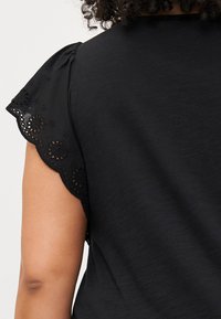 Close-up of a person’s shoulder and arm wearing a black blouse with decorative eyelet detailing on the sleeve.