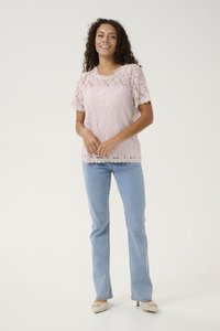 Donna sorridente con capelli ricci, che indossa un top in pizzo rosa chiaro a maniche corte, jeans azzurri e scarpe con tacco beige, in piedi su uno sfondo bianco.