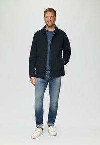Dunkelblaue Baumwolljacke mit Knopfverschluss, zwei aufgesetzten Taschen, kombiniert mit hellblauen Jeans und weißen Sneakers. Schlichter Hintergrund.