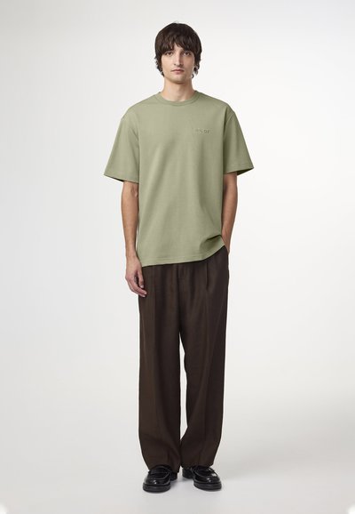 Ljust grön kortärmad t-shirt med rak skärning, i kombination med löst sittande bruna byxor och svarta skor, presenterad mot en vit bakgrund.