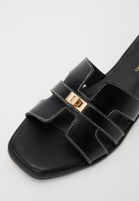 Svart skinn slidesandal med to brede stropper, gullspenne-detalj og kontrasterende hvite sømmer. Firkantet tådesign og flat såle.