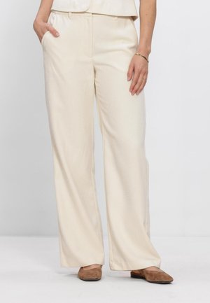 Kvinde iført cremefarvede vidde bukser og brune flade sko, stående med den ene hånd i lommen og den anden afslappet ned langs siden.