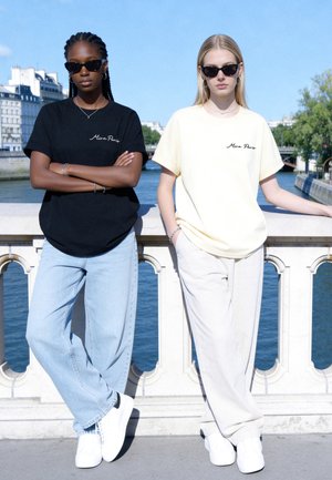 Deux femmes portant des lunettes de soleil et des t-shirts Mira Paris se tiennent près d'une rivière avec des bâtiments de la ville et un ciel clair en arrière-plan.
