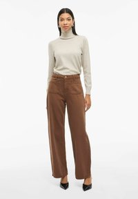 Maglione a collo alto grigio chiaro, pantaloni a gamba larga marroni con due tasche posteriori e décolleté neri a punta. Texture liscia e design moderno.