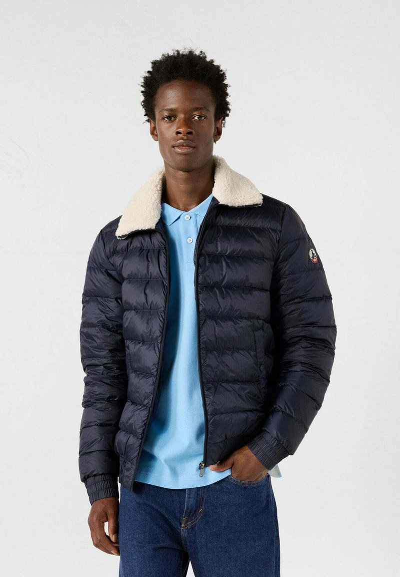 Piumino blu navy con colletto in pelliccia sintetica crema, texture trapuntata, chiusura con zip e polsini a coste. Indossato sopra una maglietta polo azzurra chiara.