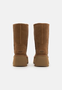 Botas de camurça castanhas até o meio da panturrilha com sola plataforma, apresentando a marca UGG na parte de trás. Textura suave e design estruturado.