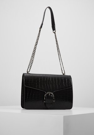 Schwarze Schultertasche aus leder mit Krokoprägung, silberner Kettenriemen und rundem Schnallverschluss, präsentiert hängend an einer weißen Fläche.