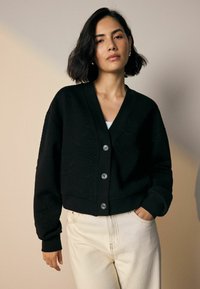 Frau mit kurzem dunklem Haar, die eine schwarze strukturierte Knopfmanschette und cremefarbene Hosen trägt, steht vor einem schlichten beigen Hintergrund.