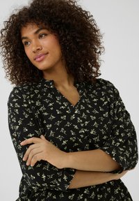 Blouse noire à manches trois quarts, ornée d'un motif floral en vert clair. Le tissu semble doux avec une coupe décontractée et un décolleté en V.