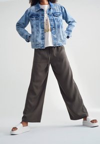  Lichtblauwe denimjack met twee voorzakken, wit graphic t-shirt, losse olive groene wijde broek en witte instap sandalen.