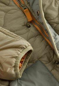 Kvadrajär beige och grå jacka med orange dragkedja och detaljer. Har elastiska manschetter och en mjuk textur. Detaljer inkluderar metallknappar.