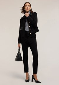 Giacca nera aderente con bottoni dorati e tasca a toppa, abbinata a un dolcevita grigio, pantaloni neri sartoriali e tacchi alti neri.