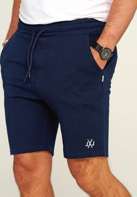Marineblaue Baumwollshorts mit einem elastischen Bund, Seitentaschen und einem weißen Logo-Akzent am Unterschenkel. Glatte Textur, knielange Passform.