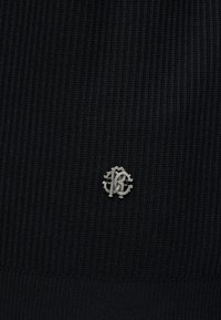 Petit logo argenté entrelacé "RB" sur tissu en maille côtelée noire, centré près du bord inférieur du textile.