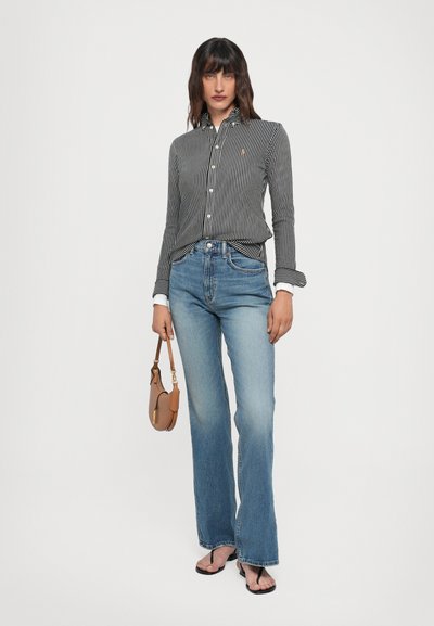 Chemise rayée noire et blanche, jean bleu légèrement évasé, tenant un sac à main marron, tongs noires, design simple à boutons.