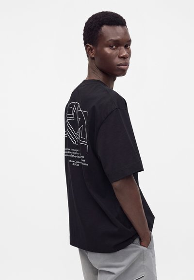 Νέος άνδρας με μαύρο γραφικό t-shirt και ανοιχτό γκρι παντελόνι στέκεται πλαγίως με τα χέρια στις τσέπες, κοιτάζοντας πάνω από τον ώμο σε απλό φόντο.