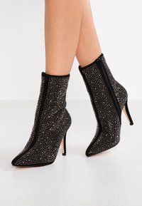 Botas de tobillo de ante negro con puntera afilada, tacones de aguja, cremalleras laterales y cubiertas de pequeños tachuelas plateadas, llevadas sobre piernas desnudas.
