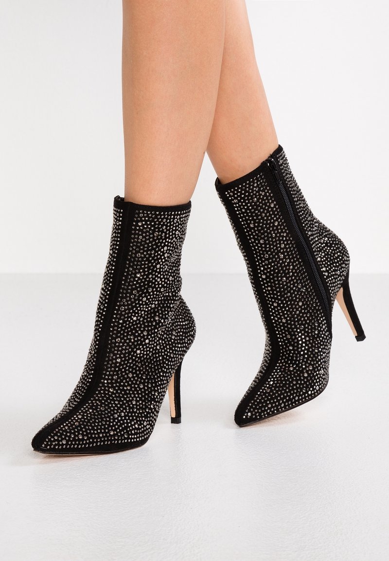 Botas de tobillo de ante negro con puntera afilada, tacones de aguja, cremalleras laterales y cubiertas de pequeños tachuelas plateadas, llevadas sobre piernas desnudas.