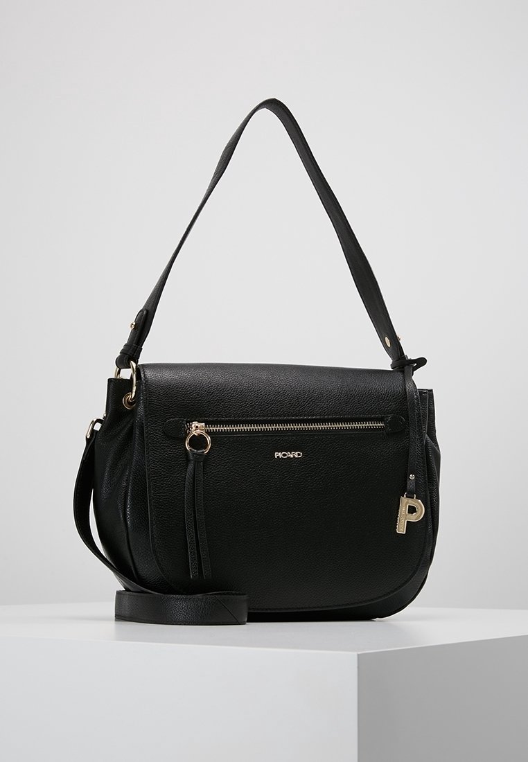 Sac à bandoulière en cuir noir avec poche avant zippée, tirette de fermeture éclair dorée, petit pendentif doré en forme de "P" et sangle ajustable, posé sur un piédestal blanc.