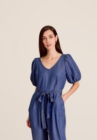 Denim jumpsuit met pofmouwen, V-hals en ceintuur in de taille. Kenmerkt zich door een diepblauwe kleur en subtiele structuur, gestyled met zijzakken.