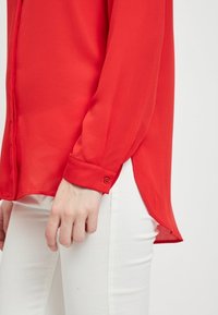 Blusa rossa trasparente con polsini abbottonati e orlo curvo. Indossata sopra pantaloni bianchi, caratterizzati da una consistenza morbida e una vestibilità ampia.