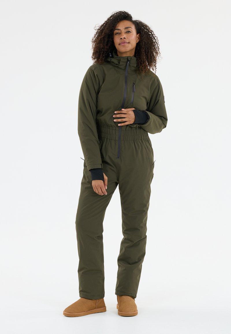 Combinaison étanche verte à col haut, avec ceinture élastique, poche zippée et accents noirs ; associée à des bottines tan à chevilles.