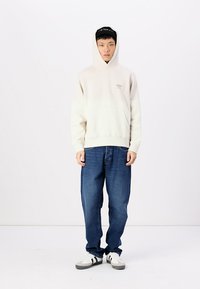 Gevoerd crèmekleurig sweatshirt met een voorzak, gedragen met iets losse blauwe jeans en witte sneakers met zwarte strepen. Eenvoudig ontwerp, geen patronen.