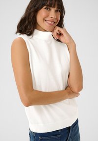 Valkoinen ilman hihoja turtleneck-toppi kevyestä, pehmeästä kankaasta. Sisältää ribsikerroksen helmassa ja hieman löysän istuvuuden. Yhdistettyinä sinisiin farkkuihin.