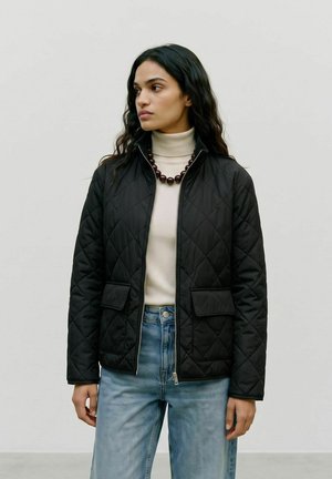 Kvinde med langt mørkt hår iført en sort quiltet jakke, cremefarvet rullekrave, blå jeans og en mørk perlekæde står foran en ensfarvet baggrund.