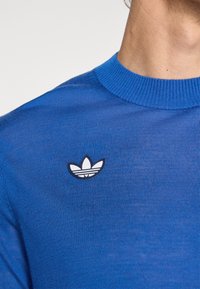 Blauwe gebreide trui met een ronde hals en een klein zwart-wit Adidas-logo. De textuur oogt glad en aansluitend.