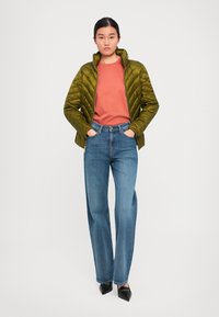 Kvinne står med hendene i lommene iført olivengrønn boblejakke, rustfarget skjorte, blå rette jeans og svarte spisse sko.