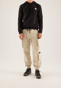 Mann trägt einen schwarzen Hoodie, beige Cargo-Jogginghosen mit Seitentaschen und schwarze Wanderschuhe und steht vor einem einfarbigen Hintergrund.