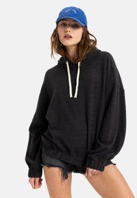 Black textured hoodie with a drawstring, paired with denim shorts. A blue cap with "ROXY 1990" logo contrasts the outfit.