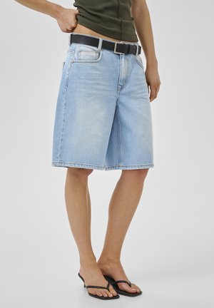 Personne portant un short en denim bleu clair longueur genou, une ceinture noire, un haut vert olive et des sandales à talons noires, debout sur fond blanc.