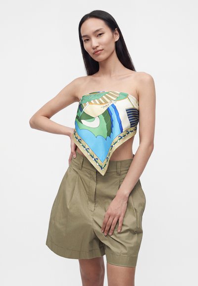Femme portant un haut bandana à motifs verts, bleus et beiges avec un short taille haute vert olive, posant avec une main sur la hanche.