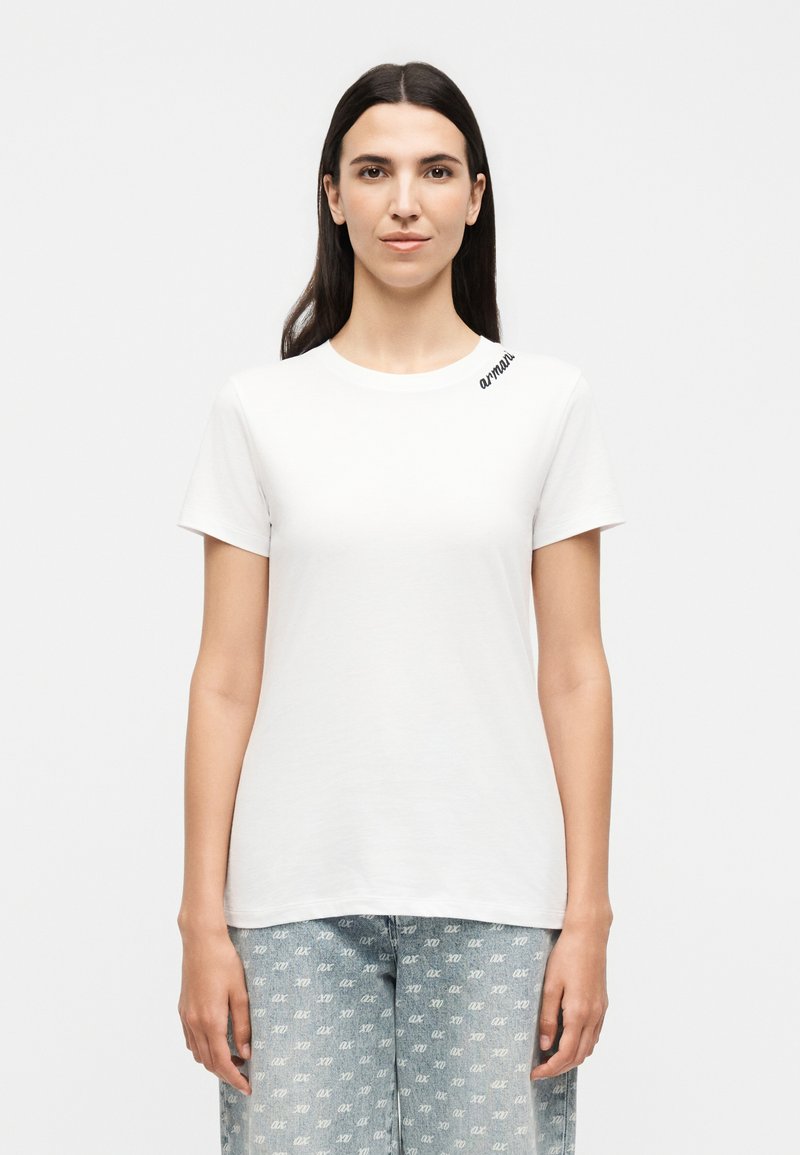 Femme aux longs cheveux foncés portant une chemise blanche à manches courtes et un pantalon bleu clair à motifs, regardant vers l'avant sur un fond uni.