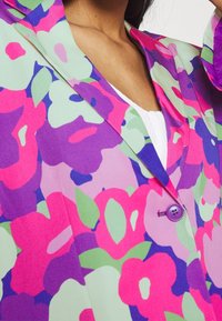 Mujer con un blazer floral colorido con botones morados sobre una camiseta blanca, mostrando el cuello y parte del brazo levantado.