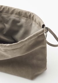 Zachte suède taupekleurige handtas met een gerimpelde bovenrand en een dunne band door een metalen ring als sluiting.
