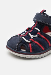 Marinblå sandal med röda och vita randiga detaljer, i mockamaterial, öppen tå-design och justerbart kardborreband. Texturerad sula.