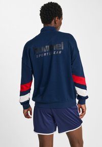 Felpa sportiva navy con colletto alto, maniche lunghe caratterizzate da accenti rossi, bianchi e blu, e un logo nero lucido sul retro.