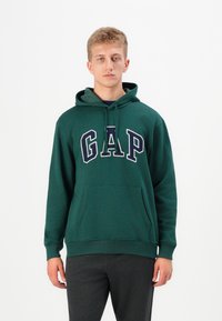 Green fleece hoodie with a kangaroo pocket, featuring large white and navy "GAP" lettering on the front. Casual fit, ribbed cuffs.
