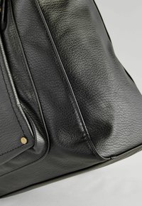 Gros plan d’un coin de sac en cuir noir texturé avec des coutures visibles et une petite rivet en laiton rond sur un fond gris clair.