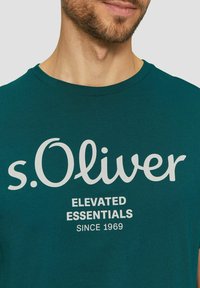 Türkisfarbiges Baumwoll-T-Shirt mit kurzen Ärmeln, das ein weißes Logo "s.Oliver" und den Text "ELEVATED ESSENTIALS SINCE 1969" in kontrastierender Schrift aufweist.