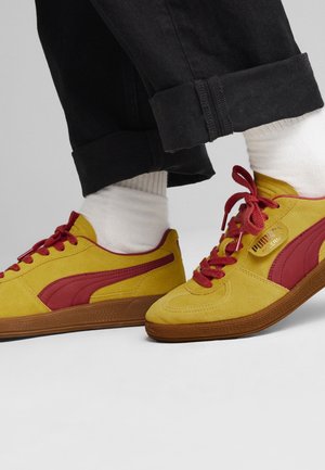 Zapatillas Puma amarillas y rojas con suelas marrones, usadas con calcetines blancos y pantalones negros remangados, sobre un fondo gris liso.