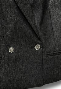 Zwarte gestructureerde blazer met een metallic glans, voorzien van twee zilveren bloemenknopen en contrasterende zakaccenten. Gepersonaliseerd en gestructureerd ontwerp.