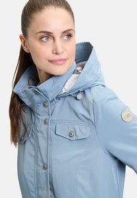 Helle blau Outdoorjacke mit Kapuze, ausgestattet mit Druckknöpfen, zwei Brusttaschen und einem Logo-Aufnäher am Ärmel. Glatte Stofftextur.