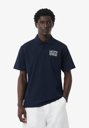 Uomo che indossa una polo Lacoste blu scuro con il logo "Roland Garros Club" e pantaloncini bianchi, in piedi davanti a uno sfondo bianco semplice.