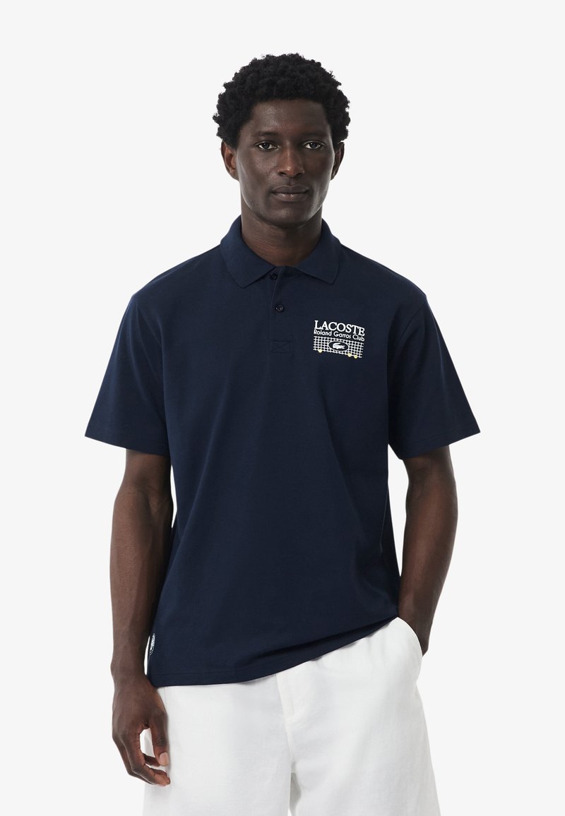 Homme portant un polo Lacoste bleu foncé avec le logo « Roland Garros Club » et un short blanc, debout devant un fond blanc uni.